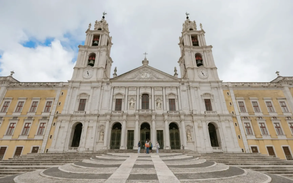 Grand staircase leading to the main entrance of the Mafra National Palace Basilica, framed by two bell towers and ornate baroque architecture.