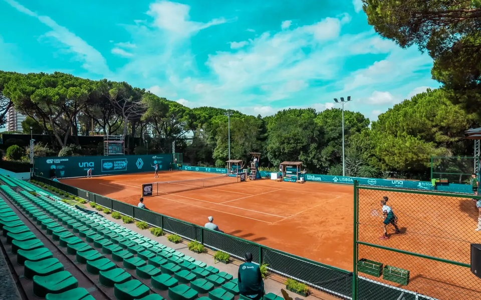 View of a clay tennis court surrounded by green bleachers and pine trees during the Lisbon Belém Open tournament, with players and officials on the field.