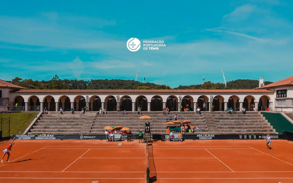 Wide view of a clay center court at the National Stadium with a tennis match in progress, flanked by stone bleachers and a colonnade building under a clear blue sky.