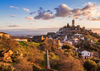 Scenic view of Monsaraz hilltop village in Alentejo, Portugal, during sunset, with stone houses, castle walls, and the Guadiana River in the background.