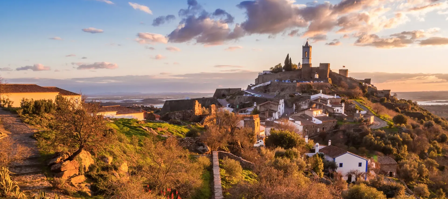 Scenic view of Monsaraz hilltop village in Alentejo, Portugal, during sunset, with stone houses, castle walls, and the Guadiana River in the background.