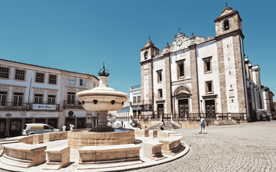 Baroque fountain and the facade of Santo Antão Church at Giraldo Square in Évora, Portugal, on a sunny day with traditional whitewashed buildings in the background.
