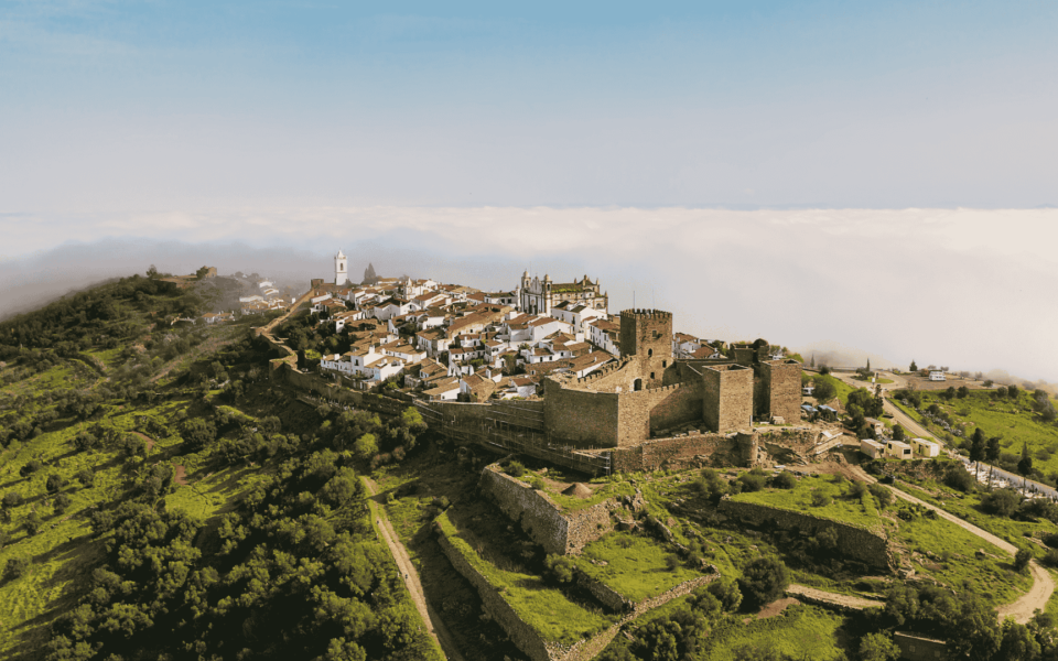 Aerial view of the walled village of Marvão and its medieval castle sitting atop a lush green mountain ridge, surrounded by low clouds in the Alentejo region of Portugal.