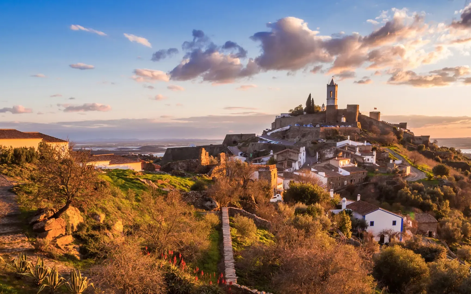 Scenic view of Monsaraz hilltop village in Alentejo, Portugal, during sunset, with stone houses, castle walls, and the Guadiana River in the background.