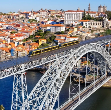 The Dom Luís I Bridge in Porto with a metro train crossing the upper deck, colorful houses of Ribeira, and the city’s historic skyline in the background.