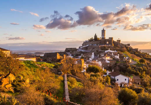 Scenic sunset view of Monsaraz, a medieval hilltop village in Alentejo, Portugal, with stone houses, castle walls, and rolling countryside lit by golden light.