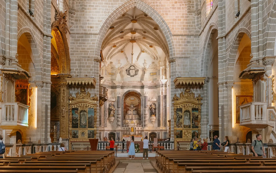 Interior of Évora Cathedral in Portugal, showcasing Gothic and Baroque architectural elements, ornate golden altarpieces, and visitors admiring the richly decorated chancel.
