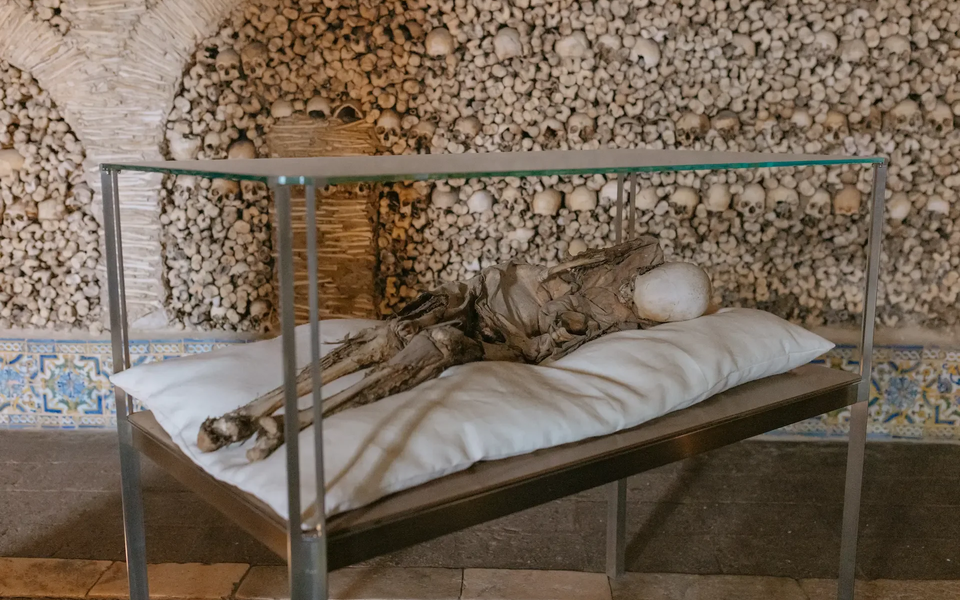 Mummified body on display inside the Chapel of Bones (Capela dos Ossos) in Évora, Portugal, with walls decorated with human skulls and bones.