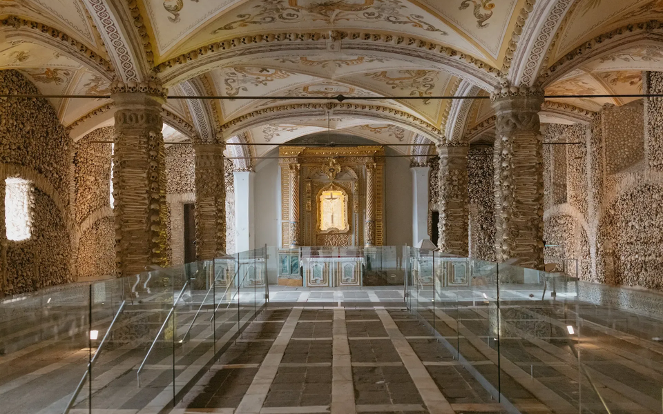 Interior view of the Chapel of Bones (Capela dos Ossos) in Évora, Portugal, showing skull-lined pillars, decorated vaulted ceilings, and a golden altar at the end of the chapel.