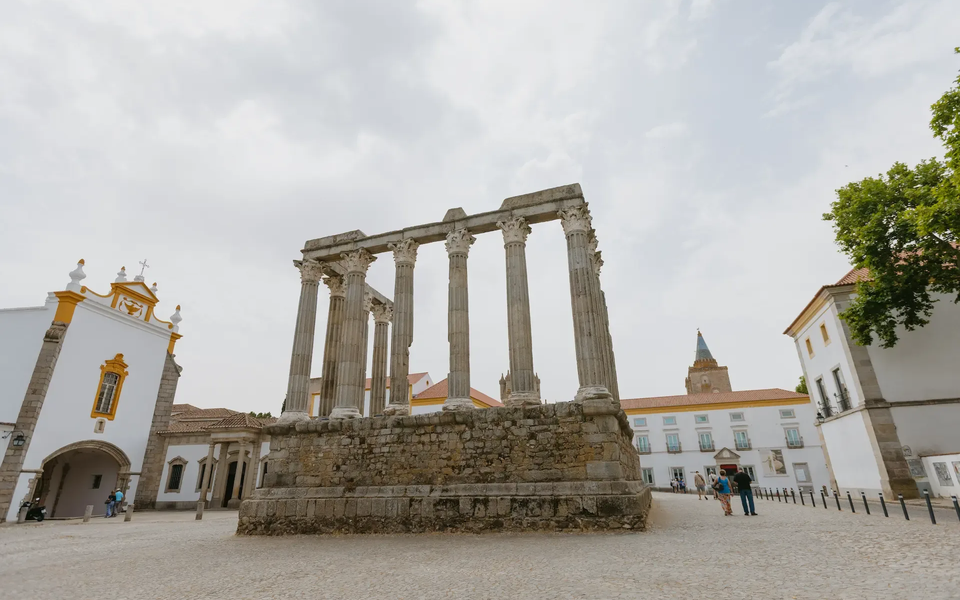 The Roman Temple of Évora, also known as the Temple of Diana, with surrounding historic buildings and tourists walking on a cobbled square in Portugal.