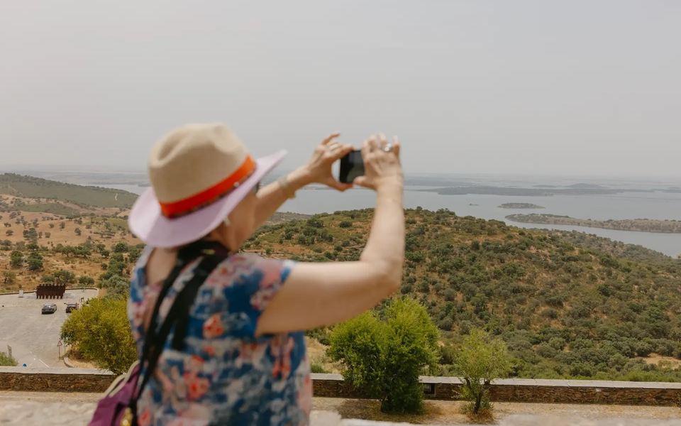 Tourist taking a photo of the panoramic view over Alqueva Lake from the medieval village of Monsaraz in Portugal, with rolling hills and calm waters in the distance.