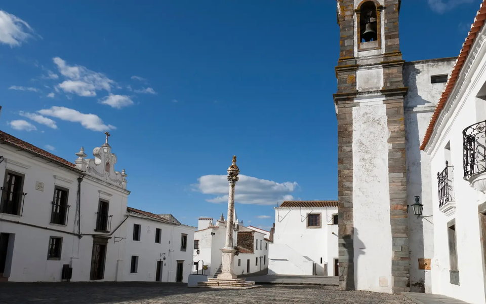 Historic main square of Monsaraz, Portugal, featuring whitewashed houses, the pillory (pelourinho), and a bell tower under a clear blue sky.