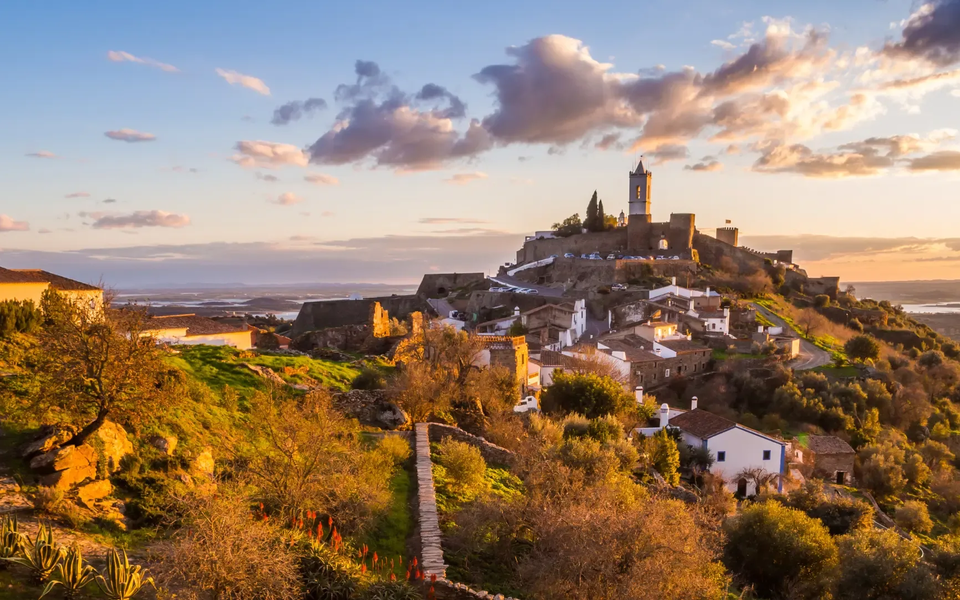 Scenic sunset view of Monsaraz, a medieval hilltop village in Alentejo, Portugal, with stone houses, castle walls, and rolling countryside lit by golden light.