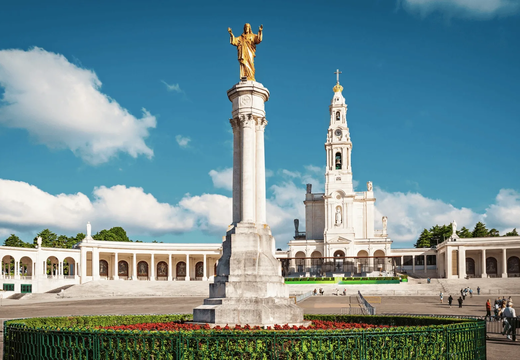 Wide-angle view of the Sanctuary of Fátima in Portugal, showing the golden statue of Jesus, the central colonnade, and the iconic basilica under a clear blue sky.