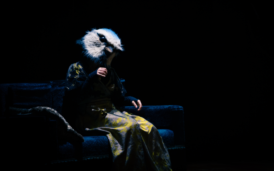 Character with a bird head mask sitting on a dark stage set