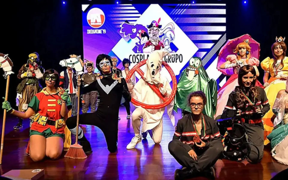 A group of cosplayers in elaborate costumes posing on stage at the Iberanime 2019 Group Cosplay Contest. The background screen displays the event logo, and the characters include references to Ghostbusters, Batman, Disney princesses, and anime.