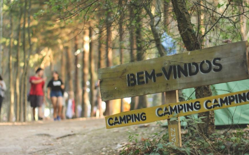 Wooden sign reading “Bem-Vindos” and “Camping” welcomes visitors to the forest campsite at Indie Music Fest 2025. In the background, people walk among tall trees and tents, evoking a relaxed, communal festival vibe.