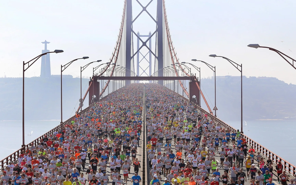 Thousands of runners crossing the 25 de Abril Bridge during a marathon in Lisbon, with the Cristo Rei statue in the background