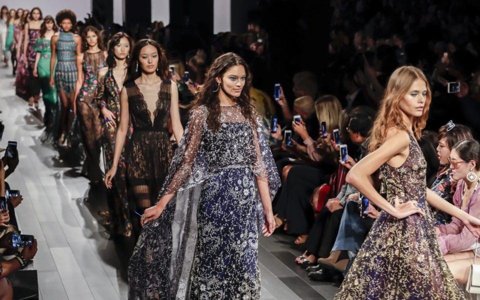 Models walking down the runway during a high-fashion show, wearing elegant embroidered dresses with the audience taking photos
