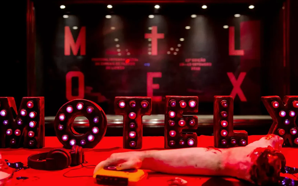 Decoração do festival MOTELX em Lisboa com luzes vermelhas intensas, letras iluminadas a formar "MOTELX", um braço falso ensanguentado e adereços de terror sobre uma mesa. Ambiente escuro e sinistro típico de um festival de cinema de terror.