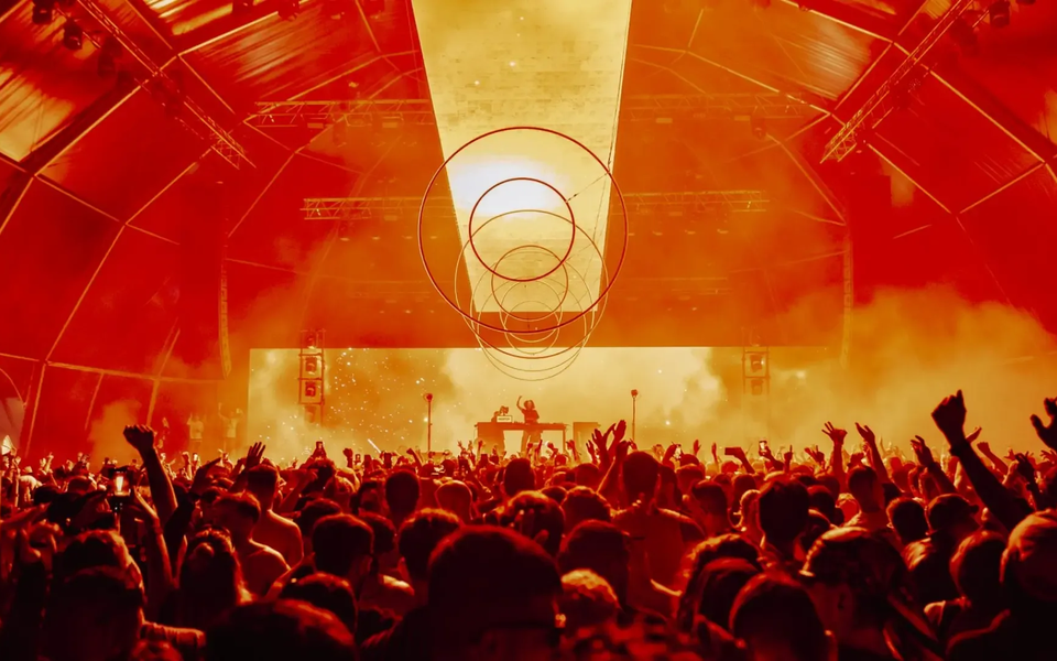 Multidão vibrante sob luzes vermelhas intensas numa tenda de festival, com DJ em palco e estrutura circular suspensa sobre o público, num concerto de música eletrónica em Lisboa.