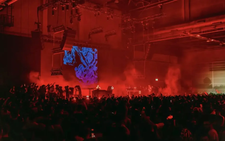 Festival de música eletrónica num armazém em Viana do Castelo, com iluminação vermelha intensa, DJ no palco e projeção digital azul psicadélica, rodeado por uma multidão vibrante.