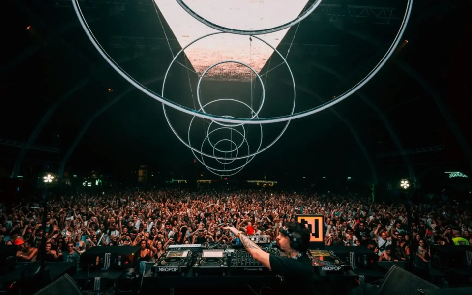 Multidão em êxtase durante atuação de DJ no Neopop Festival em Viana do Castelo, com luzes circulares suspensas e atmosfera eletrizante.