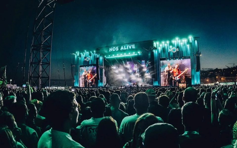 Crowd at NOS Alive Festival in Portugal during a sunset concert, with the main stage illuminated in blue and large screens showing the band