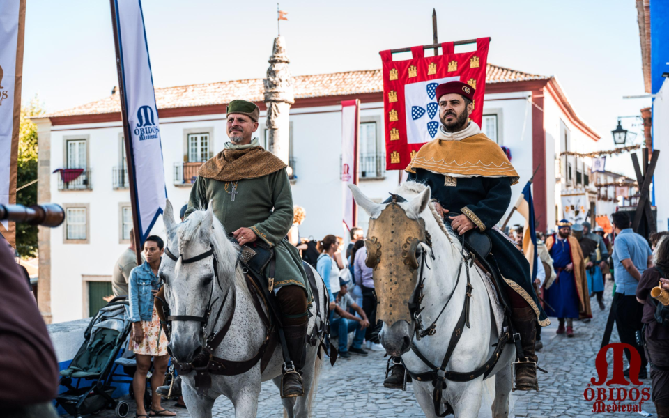 Two knights in medieval attire riding horses through the cobbled streets of Óbidos