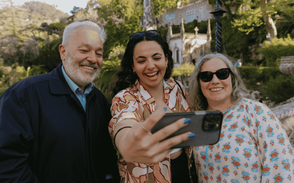 Smiling tourists taking a selfie at Quinta da Regaleira in Sintra, Portugal, with lush gardens and neo-gothic architecture in the background.