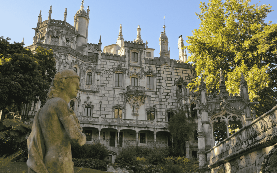 Neo-Manueline façade of the palace at Quinta da Regaleira in Sintra, Portugal, with a foreground statue and intricate stonework bathed in golden light.