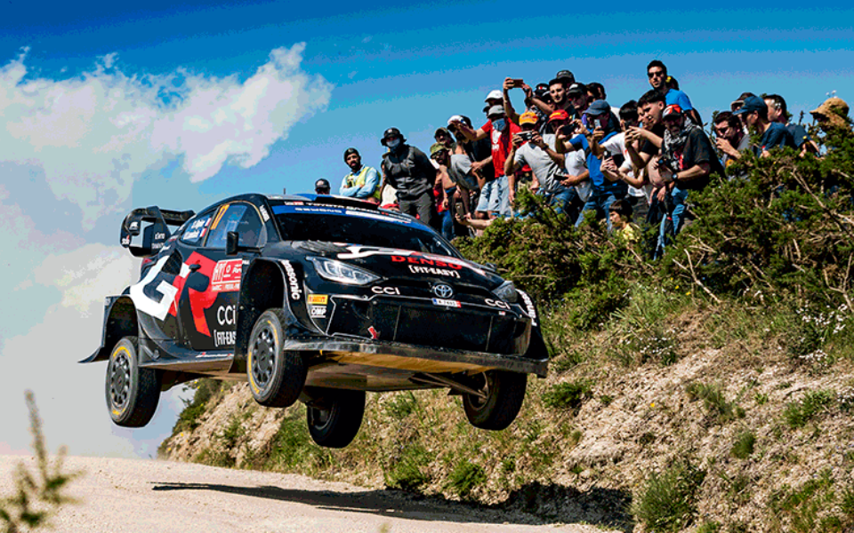 Rally car mid-air during Rally de Portugal, with enthusiastic crowd watching behind