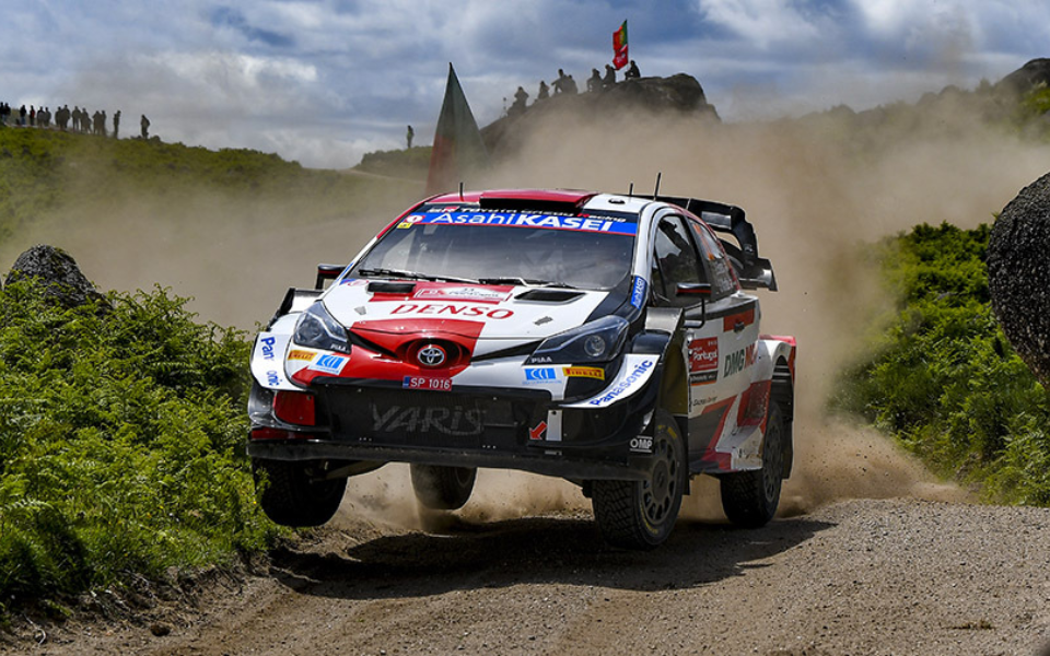 Toyota rally car turning on a dirt road surrounded by greenery, raising a cloud of dust