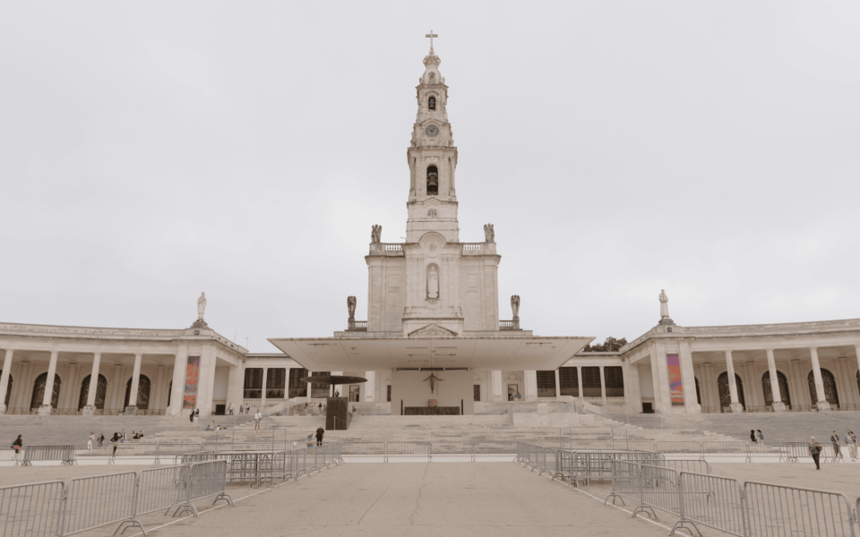 Front facade of the Basilica of Our Lady of the Rosary in Fátima, Portugal, with its bell tower and statues, under an overcast sky and surrounded by a large esplanade.