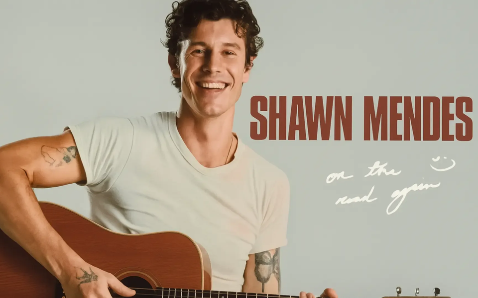 Shawn Mendes smiles while holding an acoustic guitar and wearing a white t-shirt. To his right, bold red text reads “SHAWN MENDES” with a handwritten note underneath saying “on the road again” on a light background.