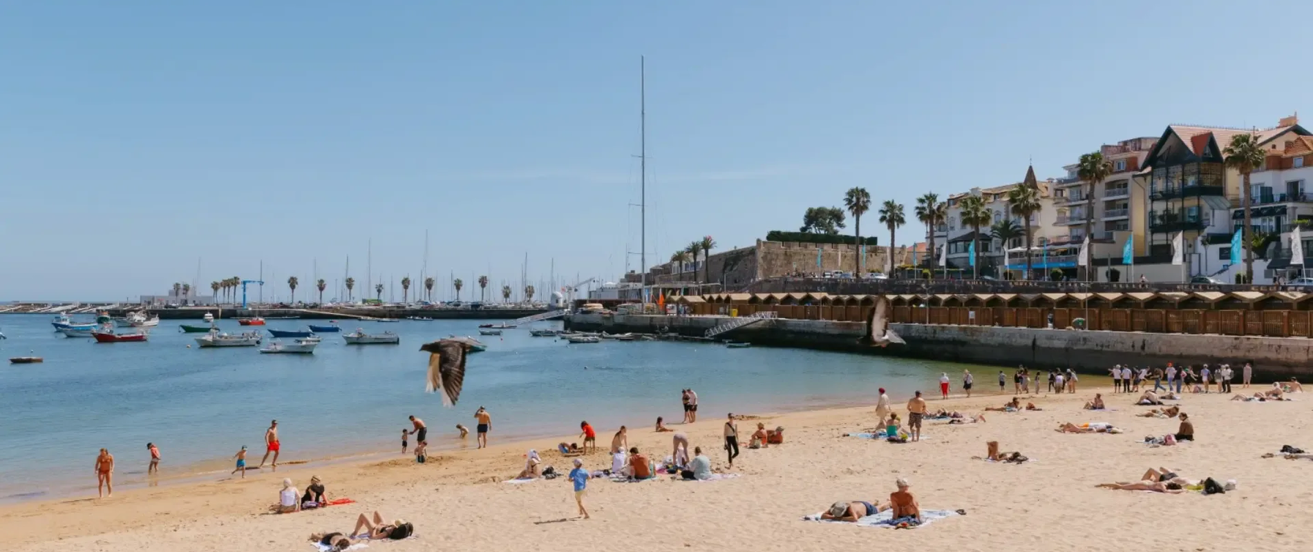 Tourists sunbathing and swimming at Praia da Ribeira beach in Cascais, Portugal, with boats anchored in the bay.