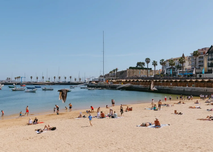 Tourists sunbathing and swimming at Praia da Ribeira beach in Cascais, Portugal, with boats anchored in the bay.