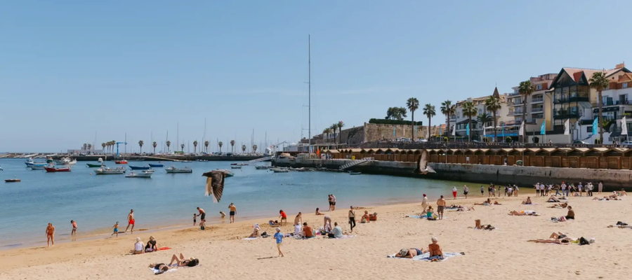 Tourists sunbathing and swimming at Praia da Ribeira beach in Cascais, Portugal, with boats anchored in the bay.