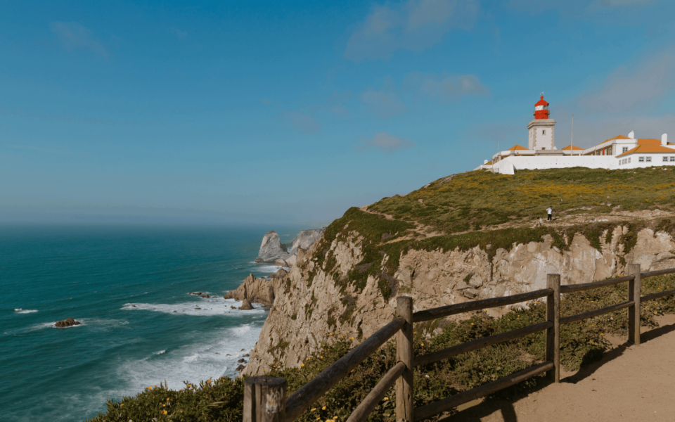 Panoramic view of the cliffs and lighthouse at Cabo da Roca, Portugal, the westernmost point of mainland Europe, with the Atlantic Ocean and rugged coastline under a blue sky.