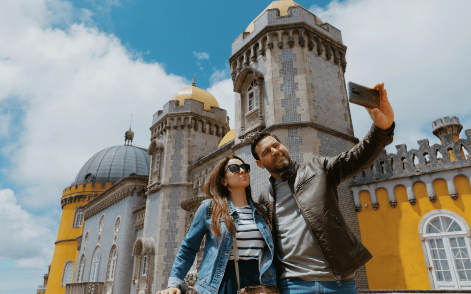 Couple taking a selfie in front of the colorful façade of Pena Palace in Sintra, Portugal, with domes and tiled towers in the background on a sunny day.