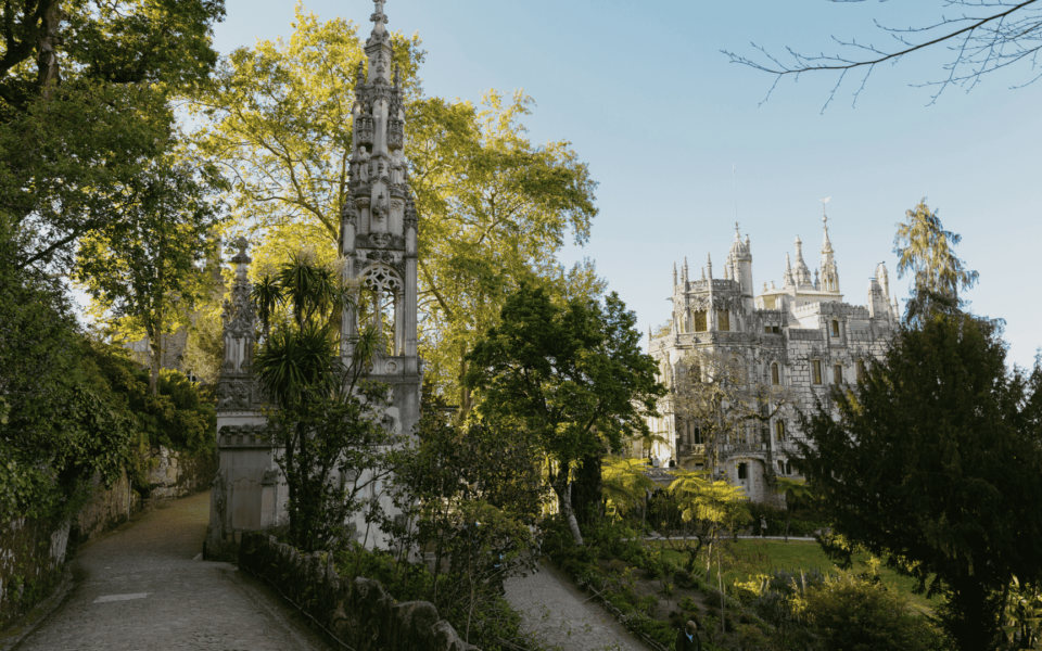Gothic tower and main palace of Quinta da Regaleira in Sintra, Portugal, surrounded by lush gardens and forest pathways on a sunny day.