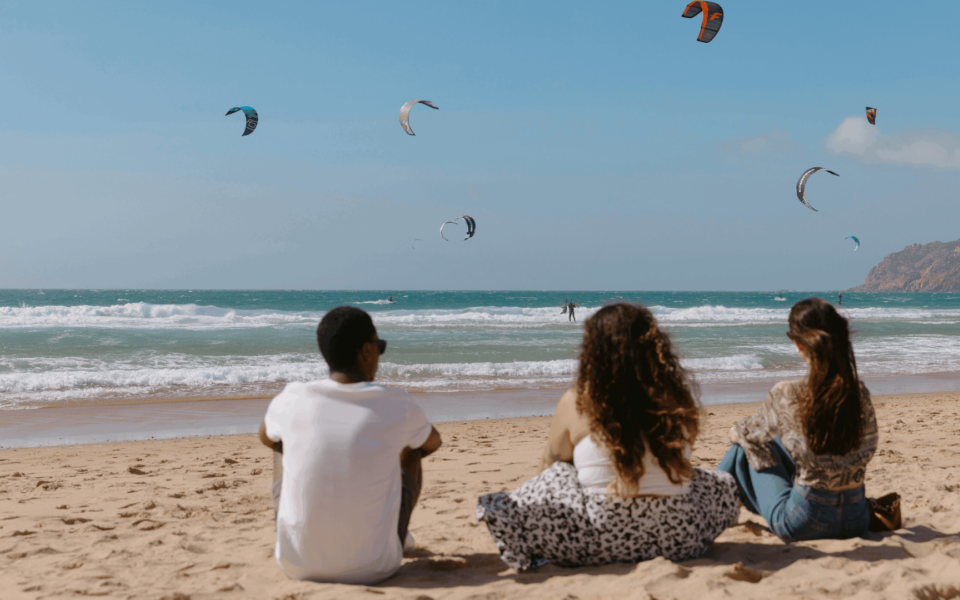 Group of friends watching kitesurfers at Guincho Beach in Cascais, Portugal, with strong waves, sandy shore, and colorful kites flying over the Atlantic Ocean.