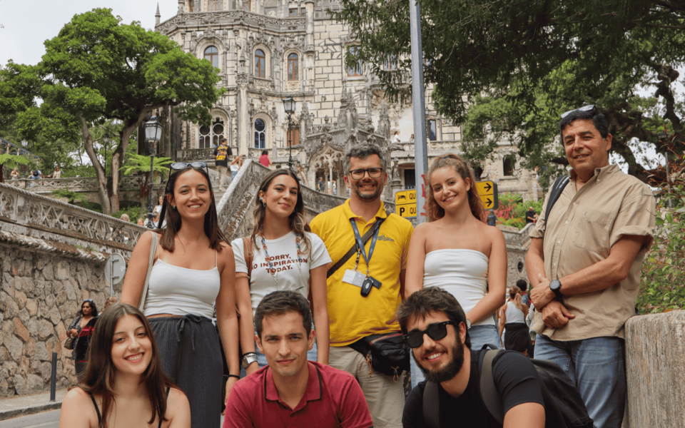 Smiling group of tourists posing at the entrance stairs of Quinta da Regaleira in Sintra, Portugal, with the ornate palace facade in the background.