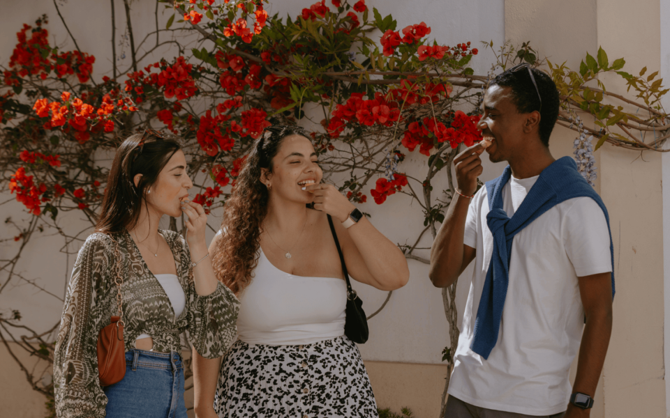 Group of friends enjoying traditional Portuguese sweets in a picturesque street with red bougainvillea flowers in the background.