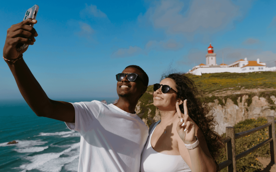 Smiling couple taking a selfie at Cabo da Roca, Portugal, with the Atlantic Ocean and the red lighthouse in the background on a sunny day.