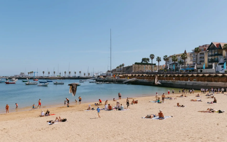 Tourists sunbathing and swimming at Praia da Ribeira beach in Cascais, Portugal, with boats anchored in the bay.
