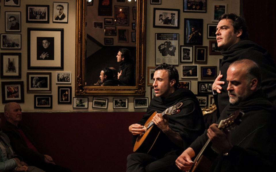 Singers and musicians wearing black cloaks perform in a fado house with photo-covered walls
