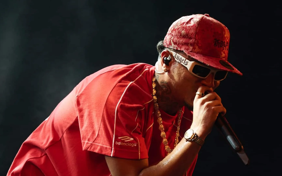 Rapper performing live on stage wearing a red Balenciaga shirt, red cap with Asian characters, white sunglasses, and gold accessories
