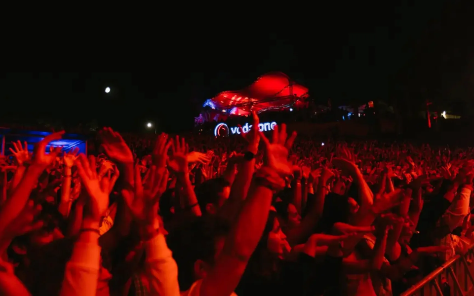 Excited crowd with hands raised at a nighttime music festival in Portugal, illuminated by red stage lights.