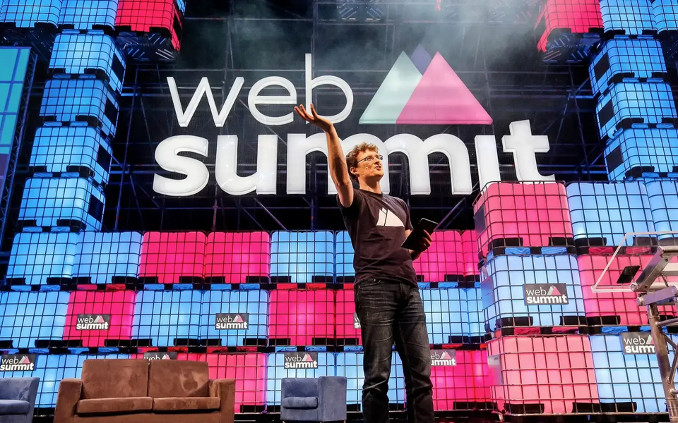 Paddy Cosgrave, co-founder of Web Summit, stands on the main stage in Lisbon, gesturing to the audience with a dynamic backdrop of pink and blue stacked cubes and the event logo.